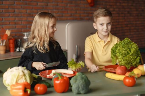 Positive little girl and boy sitting at table with assorted vegetables and fruits while preparing healthy food together in loft kitchen with brick wall