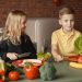Positive little girl and boy sitting at table with assorted vegetables and fruits while preparing healthy food together in loft kitchen with brick wall